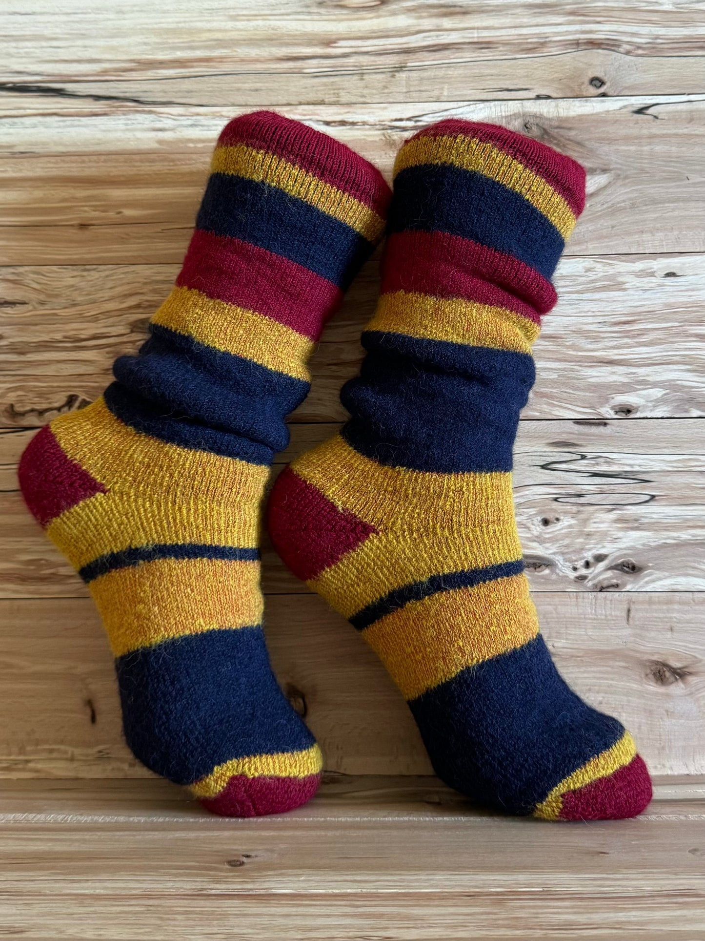 BAS THERMAL EN ALPAGA BORÉAL ENFANTS à rayures rouges, jaunes et bleues sur fond bois, parfaits pour garder les pieds au chaud en hiver. Chaussettes épaisses en fibre d'alpaga adaptées aux enfants pour un confort thermique optimal.